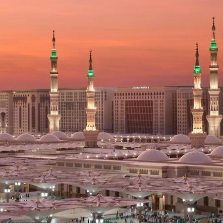Intercontinental Dar Al Iman Madinah By Ihg Medina Exterior foto The photo depicts a scenic view of a mosque complex, likely during sunset, with several minarets illuminated and showcasing green lights. The architecture features domed roofs, and the setting includes a backdrop of buildings, all bathed in a warm, o