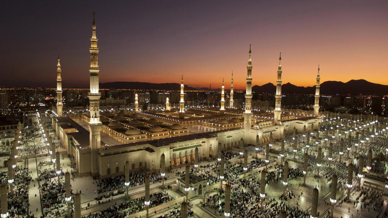 Intercontinental Dar Al Iman Madinah By Ihg Medina Exterior foto The photo depicts an aerial view of a large mosque complex during sunset. The mosque features multiple tall minarets, which are intricately designed and illuminated, giving the scene a stunning appearance as the sky transitions from daylight to dusk.