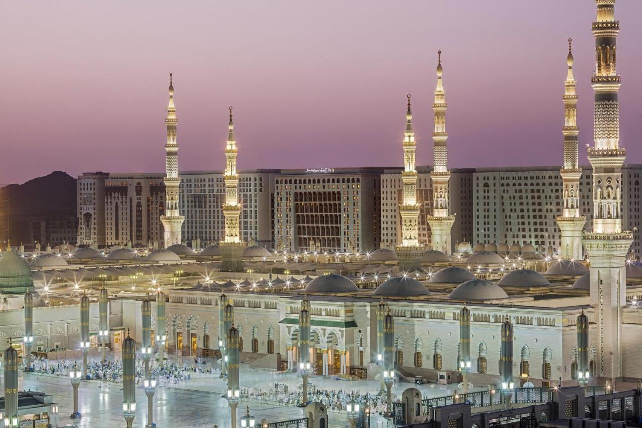 Intercontinental Dar Al Iman Madinah By Ihg Medina Exterior foto The photo shows a view of an expansive mosque complex, featuring several tall minarets that are illuminated against a twilight sky. The structure appears grand and is likely adorned with intricate architectural details. There are people gathered in t