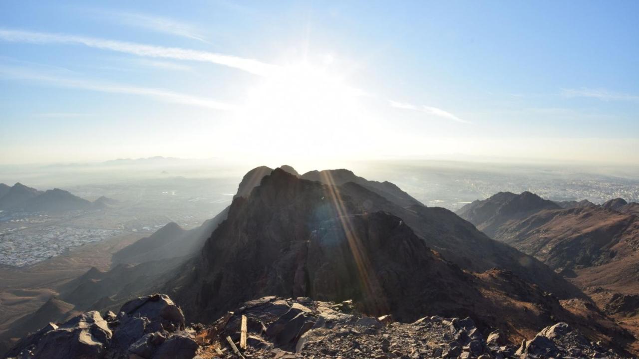 Intercontinental Dar Al Iman Madinah By Ihg Medina Exterior foto The photo shows a scenic view from a mountain top, with peaks silhouetted against the bright sun. The sun is positioned low in the sky, creating a warm glow and illuminating the landscape below. In the foreground, there are rocky terrains, while the 