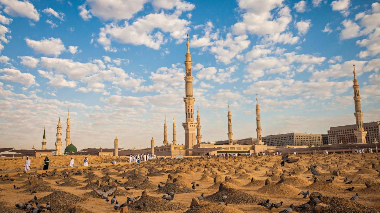 Intercontinental Dar Al Iman Madinah By Ihg Medina Exterior foto The photo shows a landscape that features a large mosque with a tall minaret surrounded by smaller minarets. In the foreground, there are numerous grave markers arranged in a desert-like setting. The sky is bright with scattered clouds, and there are