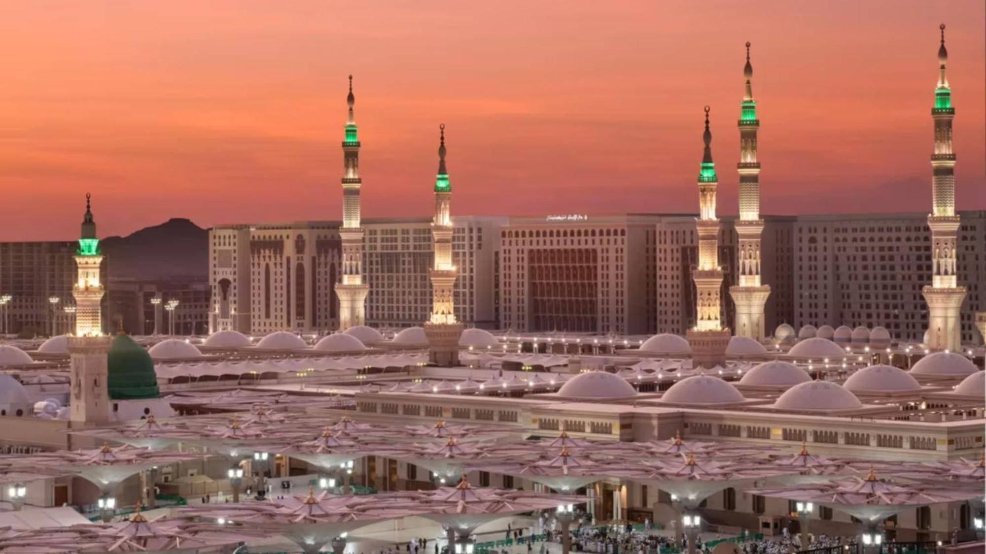 Intercontinental Dar Al Iman Madinah By Ihg Medina Exterior foto The photo depicts a scenic view of a mosque complex, likely during sunset, with several minarets illuminated and showcasing green lights. The architecture features domed roofs, and the setting includes a backdrop of buildings, all bathed in a warm, o