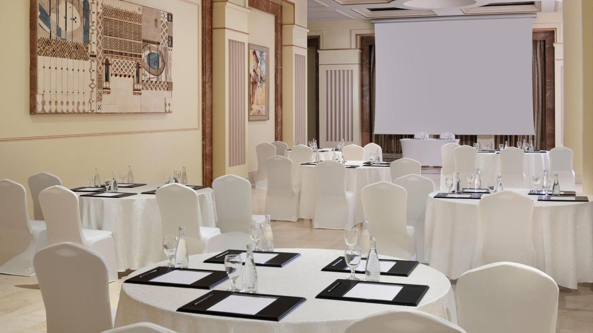 Intercontinental Dar Al Iman Madinah By Ihg Medina Exterior foto The photo depicts an elegantly arranged event space. There are several round tables covered with white tablecloths and surrounded by white chairs. Each table is set with a black folder or menu and a glass of water. The walls are adorned with artworks