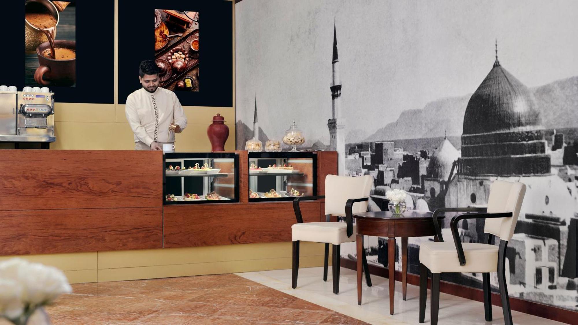 Intercontinental Dar Al Iman Madinah By Ihg Medina Exterior foto The photo shows an elegant cafe or dessert shop interior. A man in traditional attire is standing behind a wooden counter, where various pastries and sweets are displayed. The walls feature a large black-and-white mural of a cityscape, possibly depic