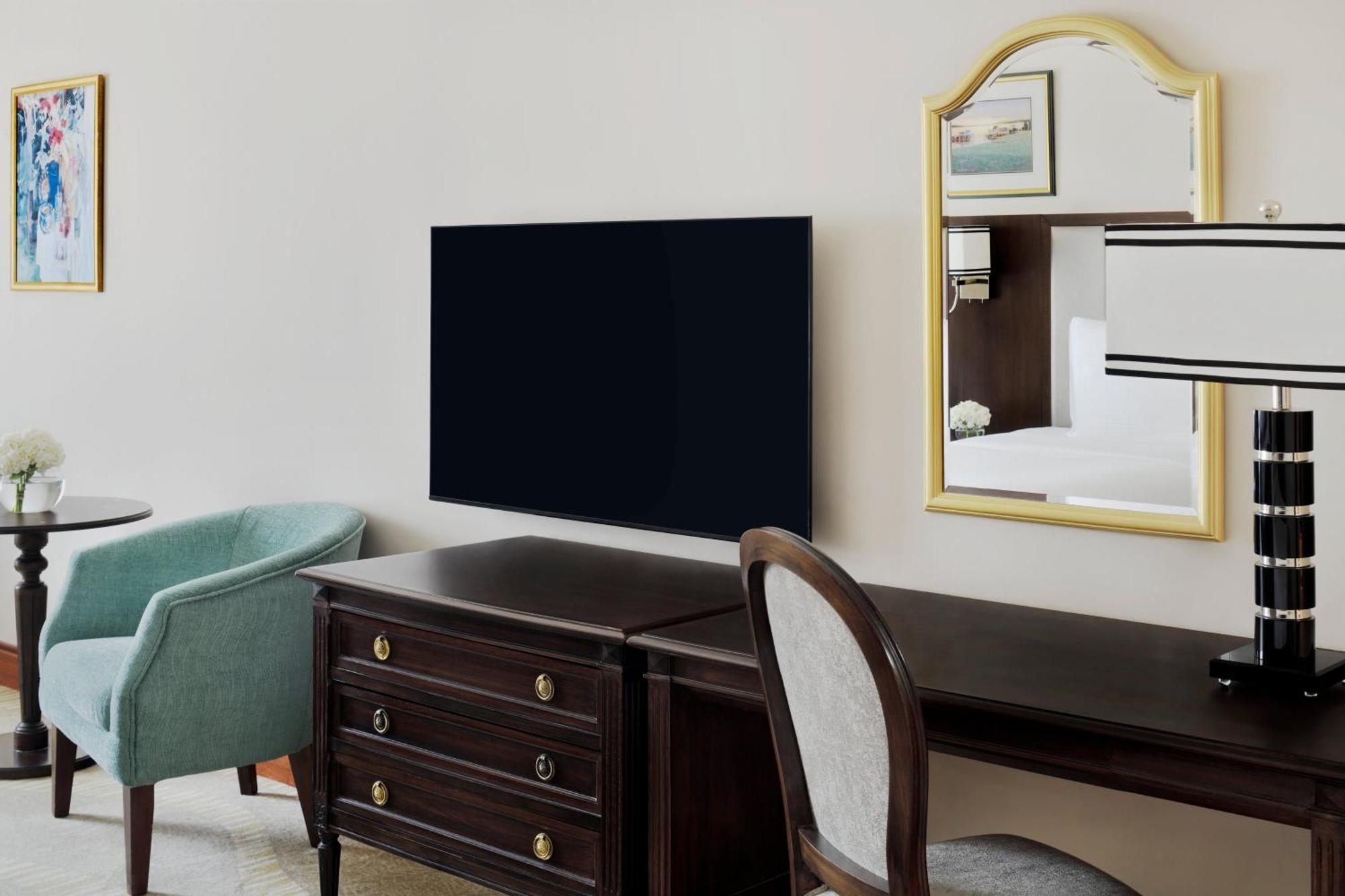 Intercontinental Dar Al Iman Madinah By Ihg Medina Exterior foto The photo shows a hotel room interior featuring a mounted flat-screen television on the wall. Below the TV, there is a dark wooden dresser with drawers. To the right, there's a wooden desk and a chair with a light-colored seat. A small table with a l