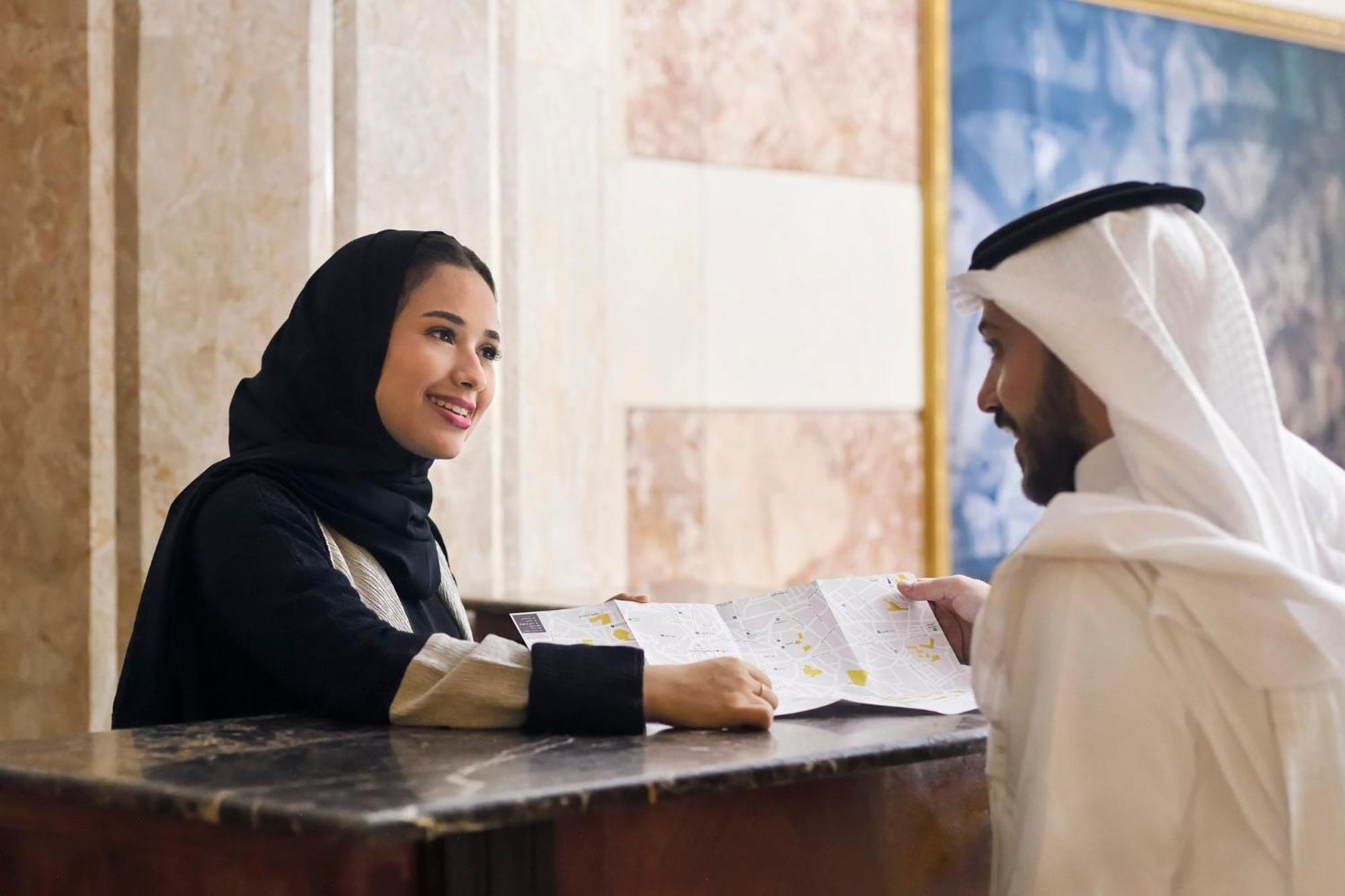 Intercontinental Dar Al Iman Madinah By Ihg Medina Exterior foto The photo depicts a woman in a black hijab and a light-colored outfit smiling as she interacts with a man wearing traditional Emirati attire, including a white kandura and ghutrah (headscarf). They are at a reception desk in what appears to be a hote