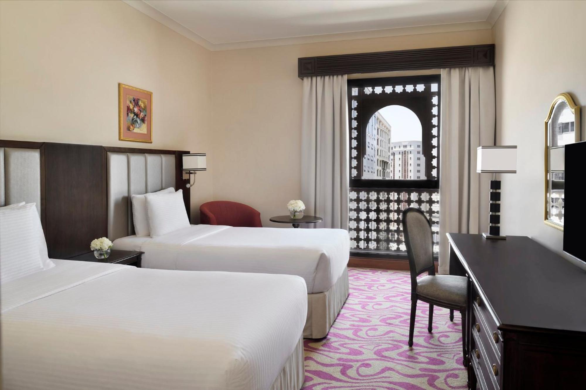 Intercontinental Dar Al Iman Madinah By Ihg Medina Exterior foto The photo shows a hotel room featuring two single beds with white bedding. The room has a light, neutral color scheme and is decorated with minimal furnishings. There's a small seating area with a red chair and a round table, as well as a desk with a