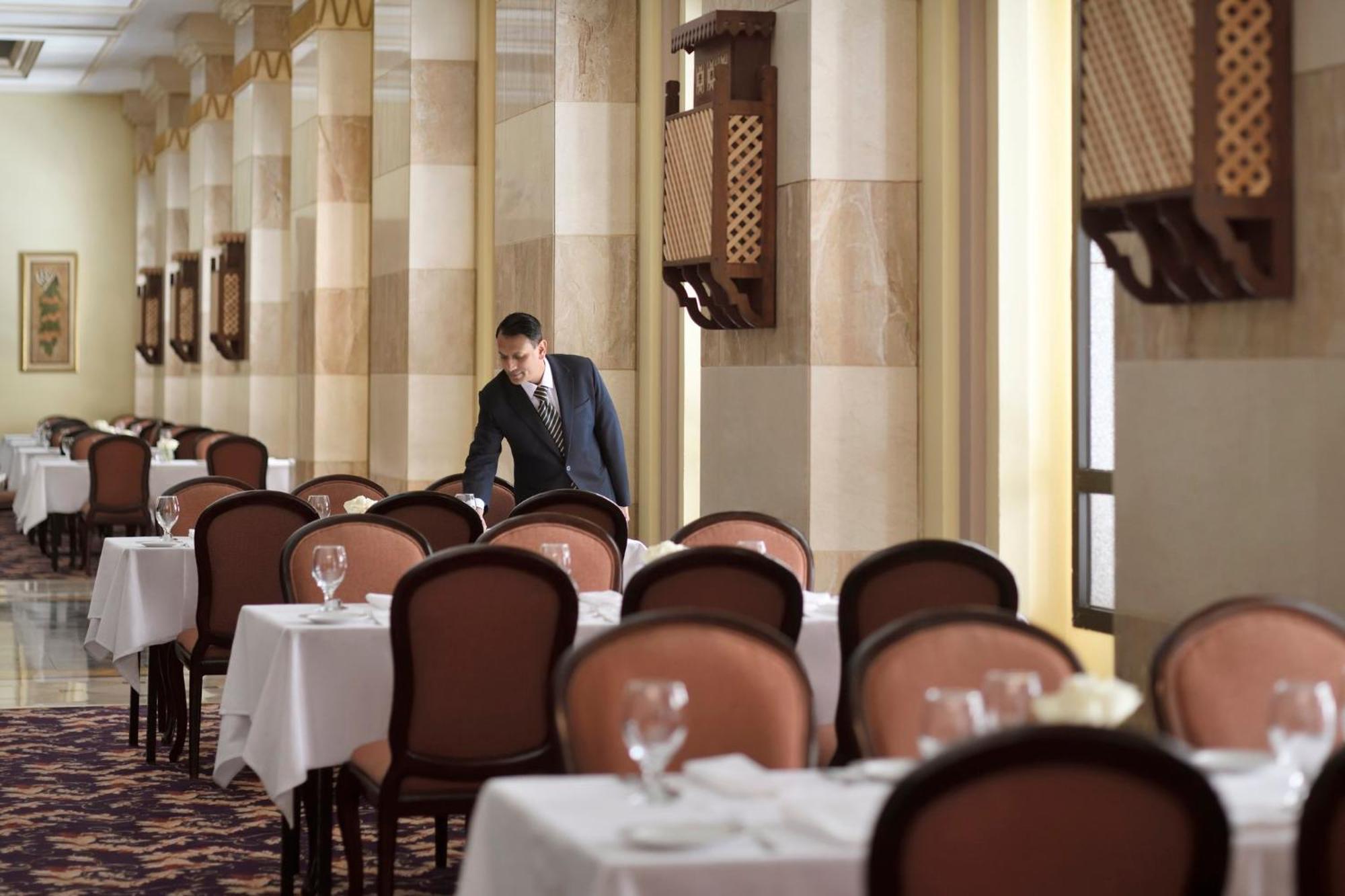 Intercontinental Dar Al Iman Madinah By Ihg Medina Exterior foto The photo depicts a dining area in an elegant restaurant. There are several tables set with white tablecloths, each accompanied by chairs. A waiter, dressed in a formal suit, is seen arranging or tending to one of the tables. The restaurant features 