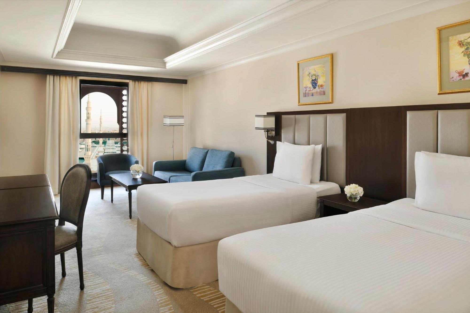 Intercontinental Dar Al Iman Madinah By Ihg Medina Exterior foto The photo shows a well-appointed hotel room featuring two twin beds with white bedding. There is a small desk with a chair, a comfortable blue sofa, and a coffee table in the sitting area. The room has a warm, neutral color scheme, accented by two pa