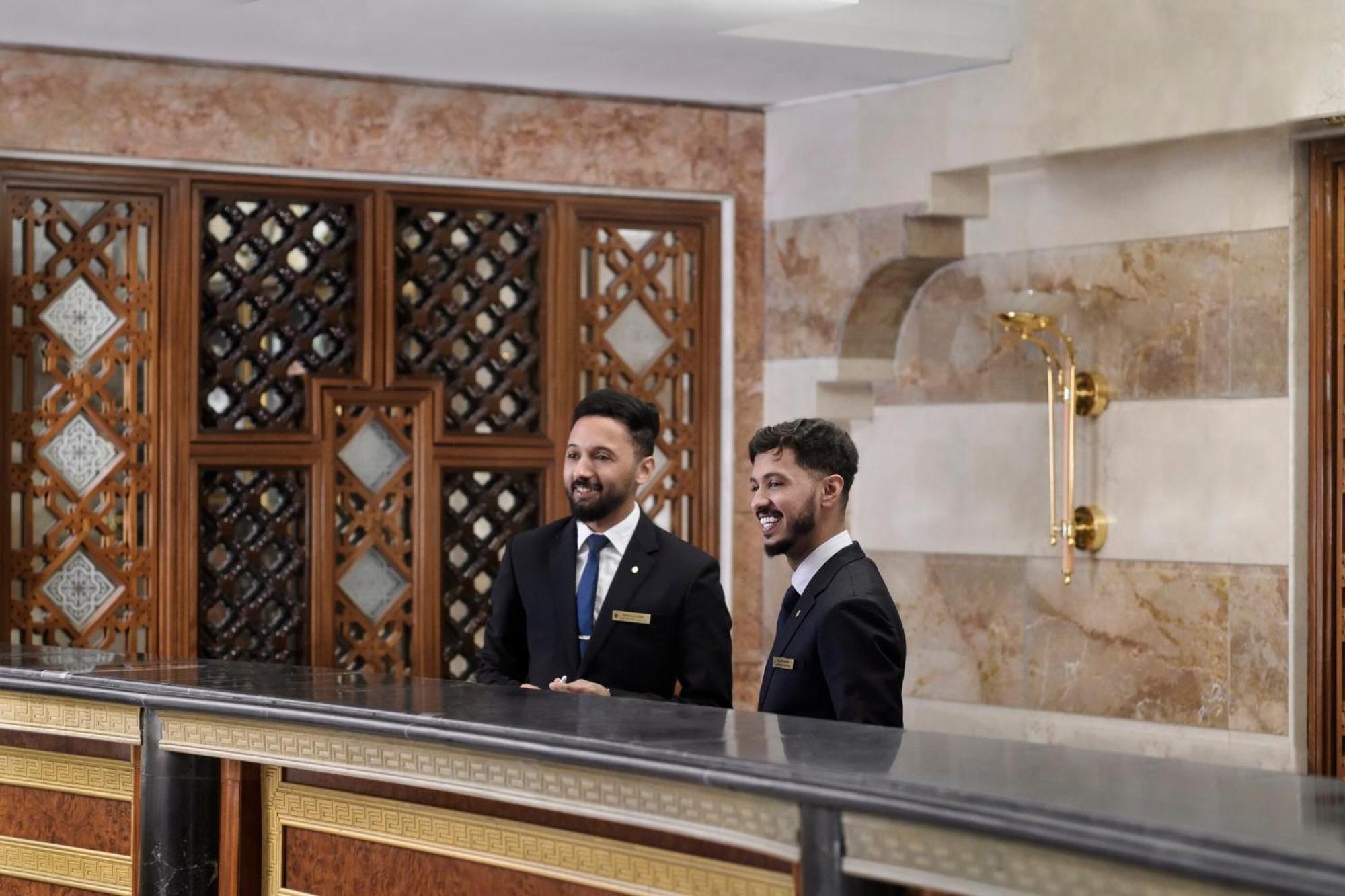 Intercontinental Dar Al Iman Madinah By Ihg Medina Exterior foto The photo shows two men standing behind a reception desk in an elegant setting, likely a hotel or similar establishment. They are both dressed in formal attire, wearing suits and ties, and appear to be smiling or engaging in conversation. The backgro