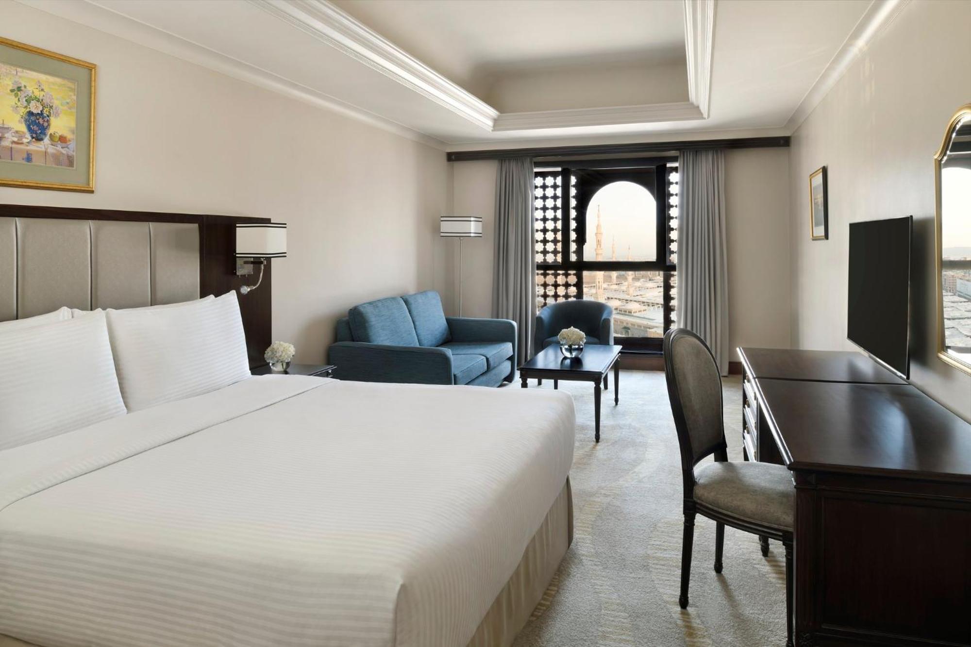 Intercontinental Dar Al Iman Madinah By Ihg Medina Exterior foto The photo shows a hotel room designed with a modern and comfortable aesthetic. It features a large bed with white linens, a seating area with a blue sofa, and a small table. There is a wooden desk with a chair and a television opposite the bed. Natur