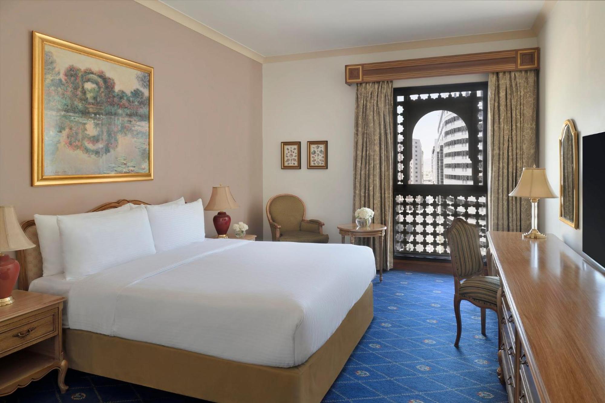 Intercontinental Dar Al Iman Madinah By Ihg Medina Exterior foto The photo shows a hotel room that features a large, neatly made bed with white linens. There is a framed painting on the wall above the bed. To the side, a small seating area includes a chair and a side table. Opposite the bed, there is a wooden desk