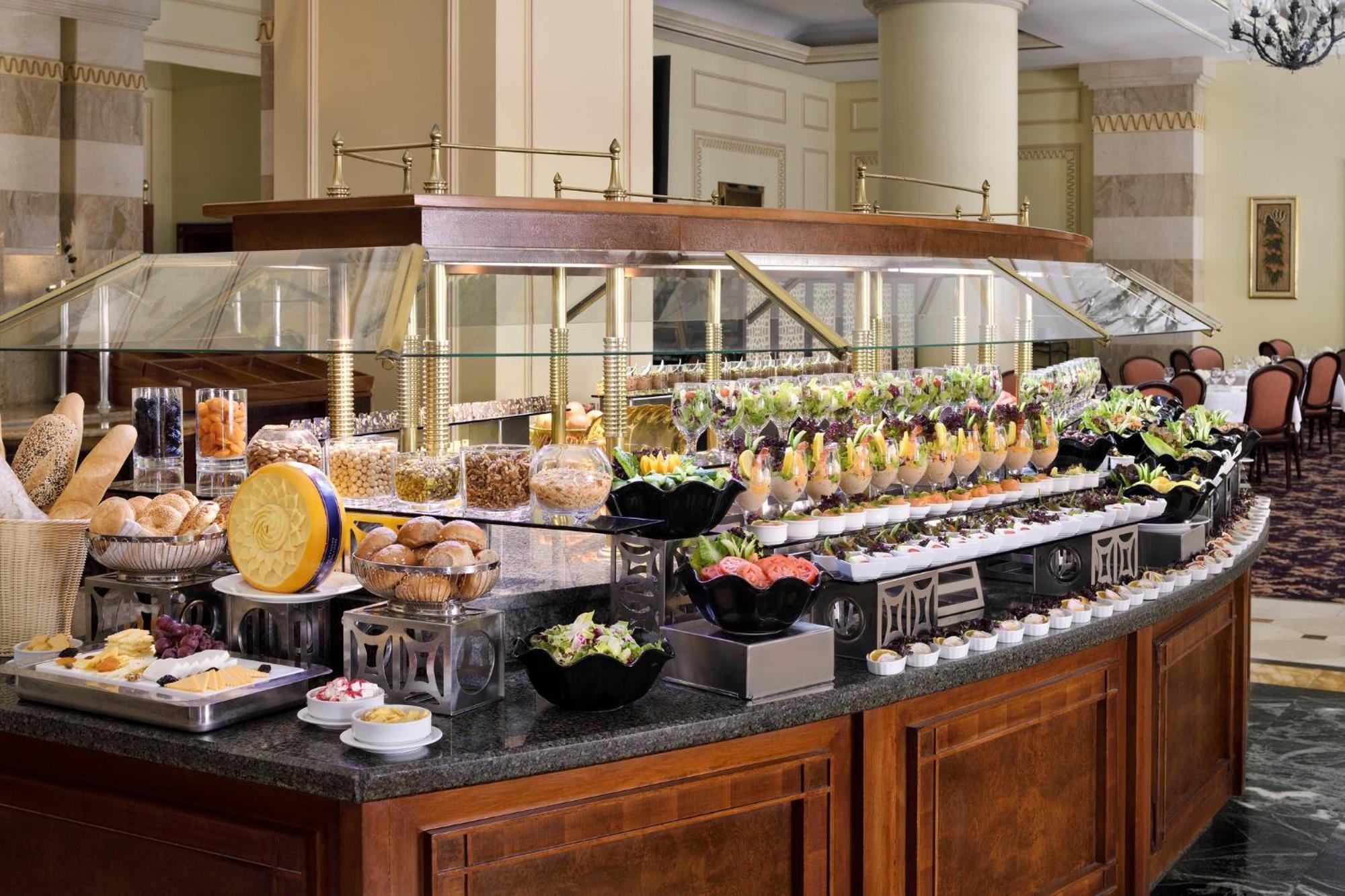 Intercontinental Dar Al Iman Madinah By Ihg Medina Exterior foto The photo shows an elegantly arranged buffet with a variety of food items. There is a large, curved wooden display with glass covers, showcasing an assortment of dishes. These include different types of breads and rolls, salads, garnishes, and desser