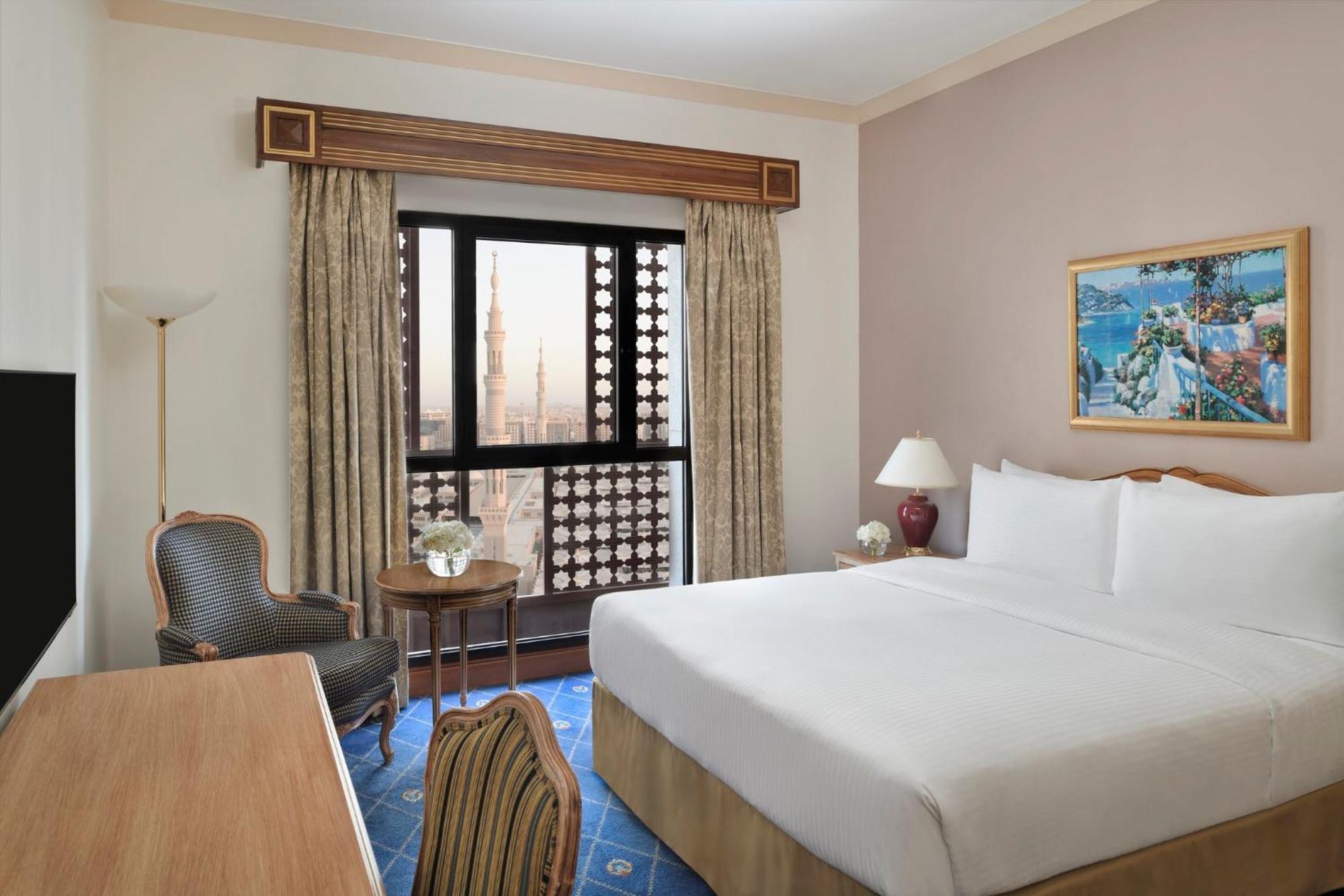 Intercontinental Dar Al Iman Madinah By Ihg Medina Exterior foto The photo shows a hotel room with a modern design. There is a large bed with white linens placed against the wall. Next to the bed, there is a small bedside table with a lamp. A comfortable armchair is situated nearby, accompanied by a wooden desk. T