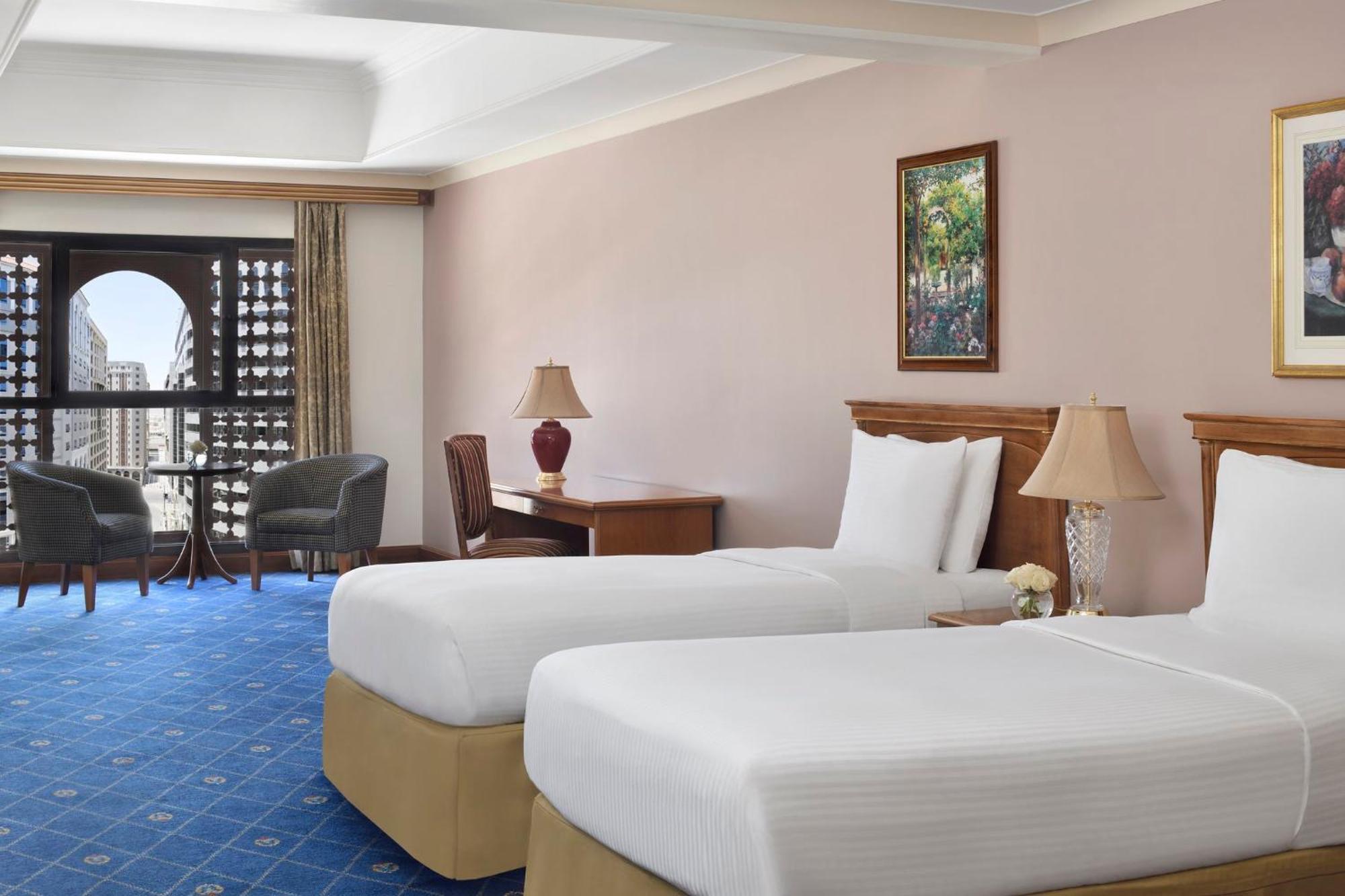 Intercontinental Dar Al Iman Madinah By Ihg Medina Exterior foto The photo shows a nicely designed hotel room featuring two twin beds with white linens and beige bases. In the background, there's a wooden desk and a lamp, along with a pair of comfortable armchairs positioned near a large window. The window showcas