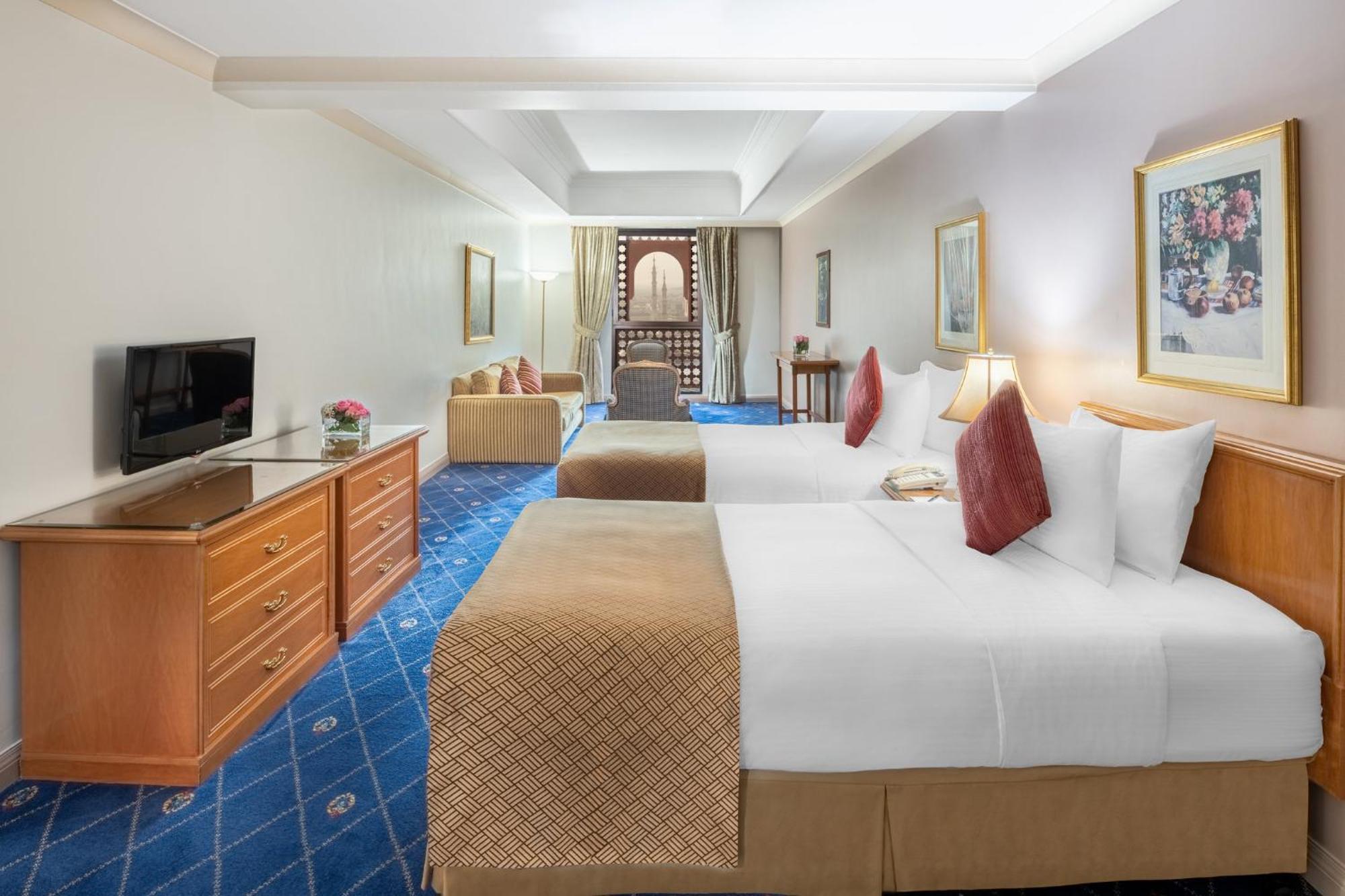 Intercontinental Dar Al Iman Madinah By Ihg Medina Exterior foto The photo shows a hotel room featuring two beds with white linens and decorative pillows. There is a wooden dresser with a television on it. The flooring has a blue carpet with a pattern, and the walls are painted in a light color. The room is well-l