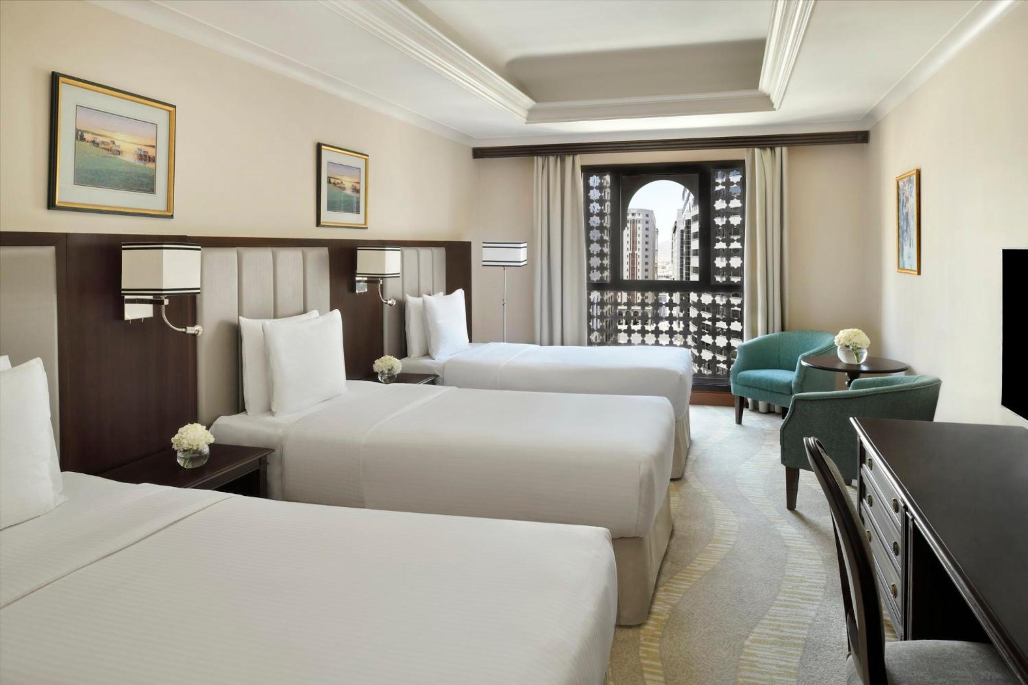 Intercontinental Dar Al Iman Madinah By Ihg Medina Exterior foto The photo shows a hotel room with three single beds, dressed in white linens. There are two paintings on the walls, depicting serene landscapes. A large window allows natural light to enter, and it is adorned with elegant curtains. A smaller seating 