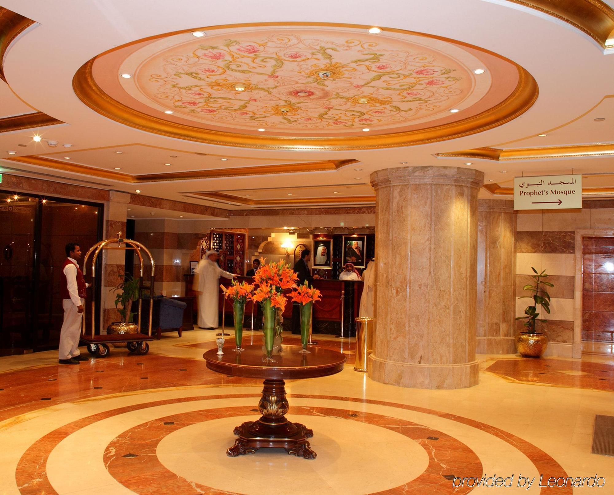 Intercontinental Dar Al Iman Madinah By Ihg Medina Interior foto The photo depicts a luxurious hotel lobby. The ceiling features an ornate design with floral patterns, adding elegance to the space. In the center of the lobby, there is a decorative table with a bowl or vase of bright orange flowers. The flooring is