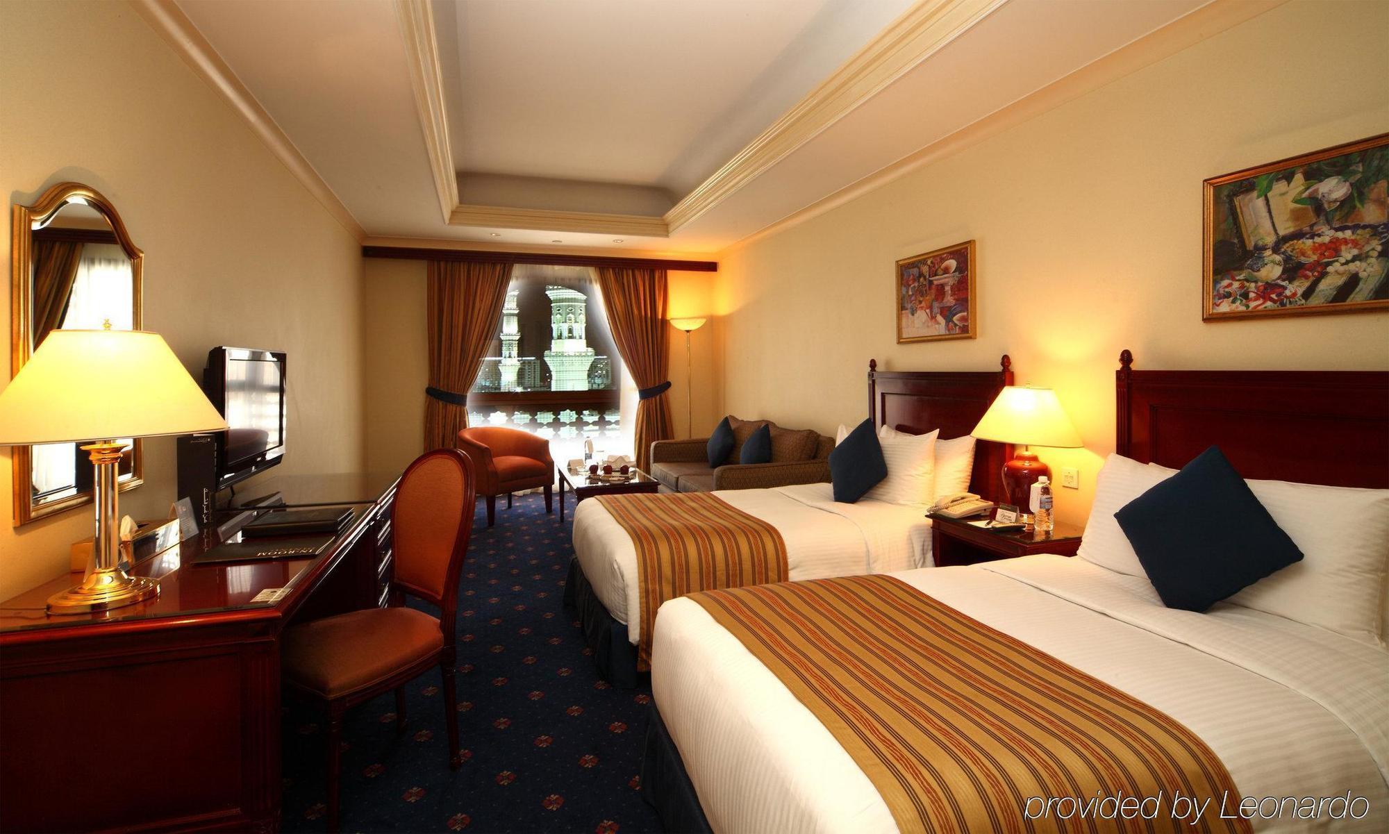 Intercontinental Dar Al Iman Madinah By Ihg Medina Quarto foto The photo shows a hotel room featuring two beds with white linens and brown striped throws. There is a wooden desk and chair, alongside a television. The walls are painted in a warm tone, and there are decorative paintings. Large windows with curtain