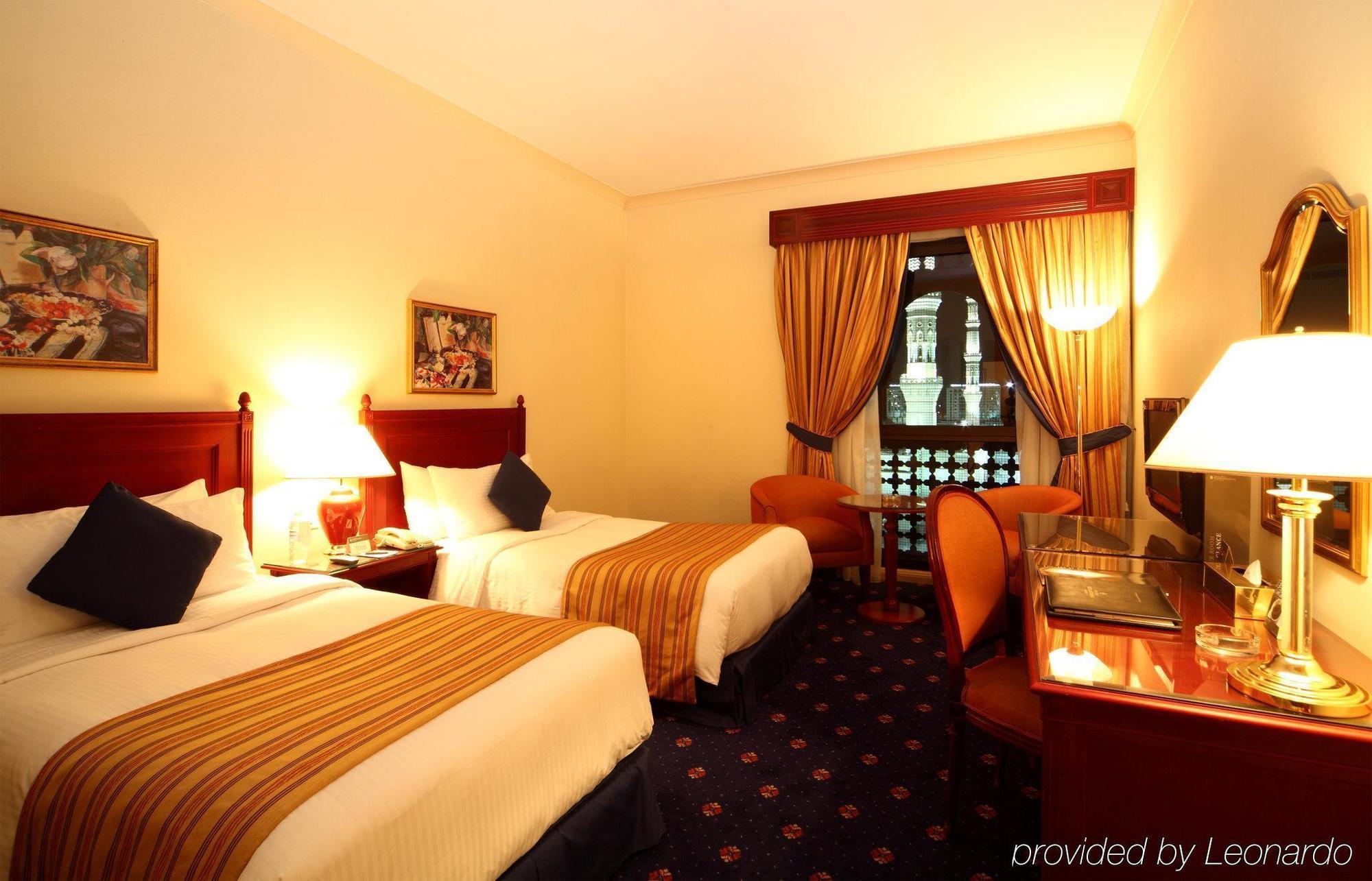 Intercontinental Dar Al Iman Madinah By Ihg Medina Quarto foto The photo shows a hotel room featuring two twin beds with white linens and brown striped bedspreads. There are decorative pillows on the beds, and the room has warm-colored walls. To the side, there's a small sitting area with a round table and a cha