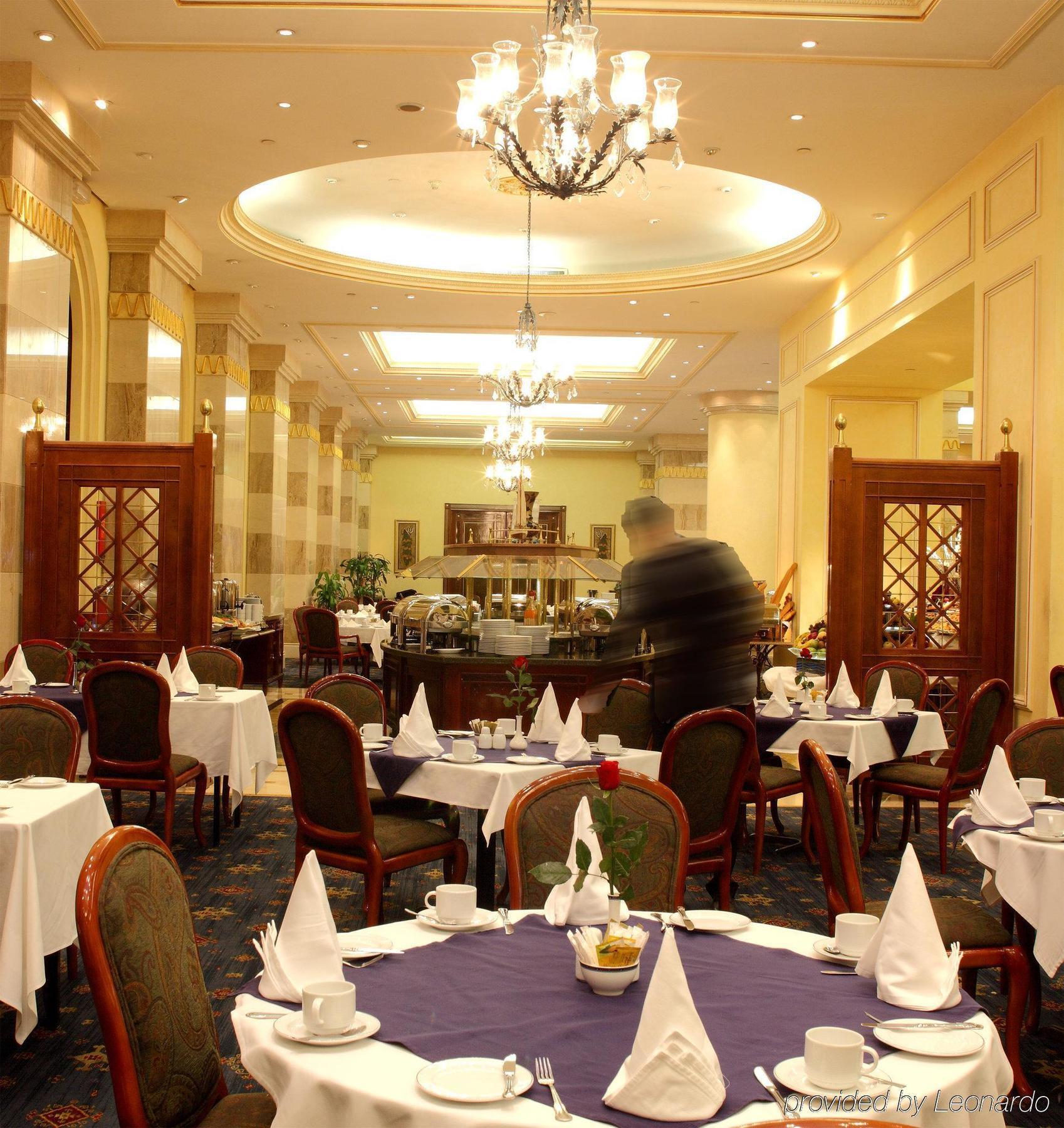Intercontinental Dar Al Iman Madinah By Ihg Medina Restaurante foto The photo depicts an elegant restaurant interior. It features round tables covered with white tablecloths, each set with plates, cutlery, and folded napkins arranged into triangles. The space has a warm, inviting atmosphere with a mix of decorative e