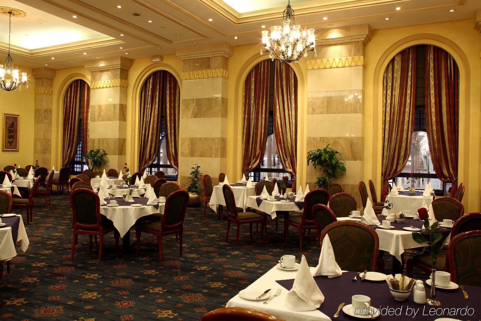Intercontinental Dar Al Iman Madinah By Ihg Medina Restaurante foto The photo depicts an elegant restaurant interior. The space features ornate decor with tall windows draped in rich, patterned curtains. Tables are neatly set with white tablecloths and napkins, and there are plush chairs arranged around them. The flo