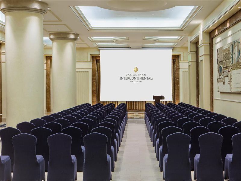 Intercontinental Dar Al Iman Madinah By Ihg Medina Exterior foto The photo shows a conference or meeting room set up. There are rows of dark blue chairs arranged facing a large screen at the front of the room. The screen displays the logo of the Intercontinental hotel, indicating that this space is likely intended