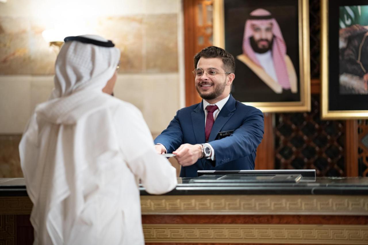 Intercontinental Dar Al Iman Madinah By Ihg Medina Exterior foto The photo shows a hotel reception area. A male receptionist, dressed in a navy suit with a tie, is smiling and handing an item to a guest who is wearing traditional white attire, possibly a kandura or thobe. The background features elegantly decorate