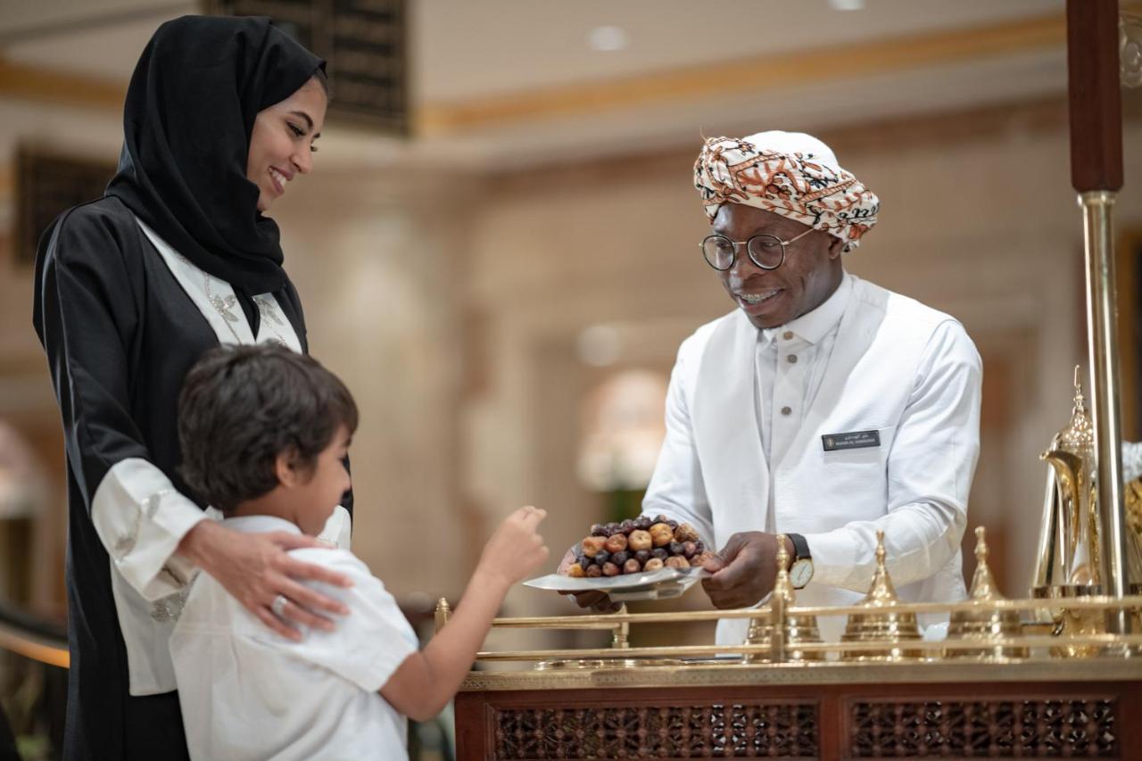 Intercontinental Dar Al Iman Madinah By Ihg Medina Exterior foto The photo depicts a warm interaction between three people. A woman, dressed in a traditional black attire and a headscarf, stands beside a young boy who is reaching out towards a plate of dates held by a smiling man in a white traditional outfit with